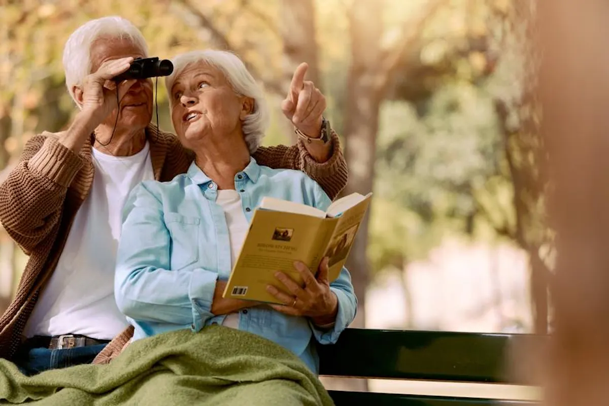 An elderly couple sits on a park bench in autumn, with the man looking through binoculars while the woman points into the distance and holds an open book.