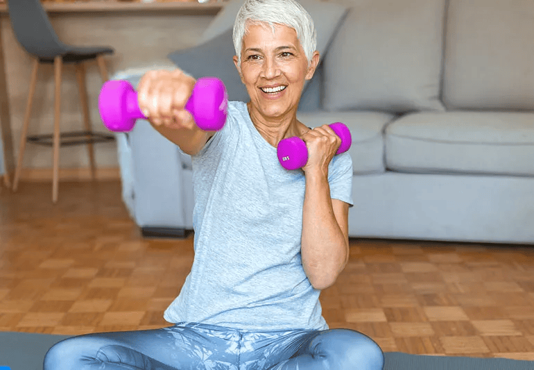 An older woman smiles while doing strength-training exercises with pink dumbbells on a yoga mat in her living room.