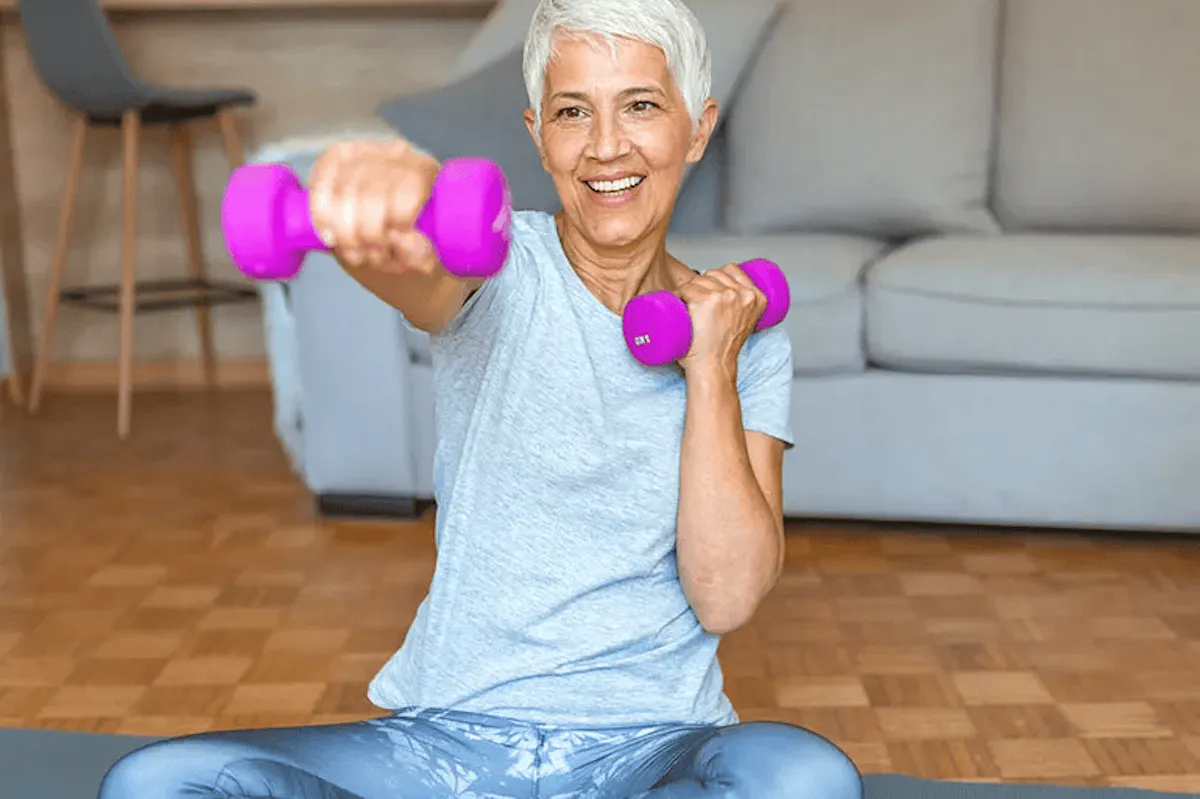 An older woman smiles while doing strength-training exercises with pink dumbbells on a yoga mat in her living room.