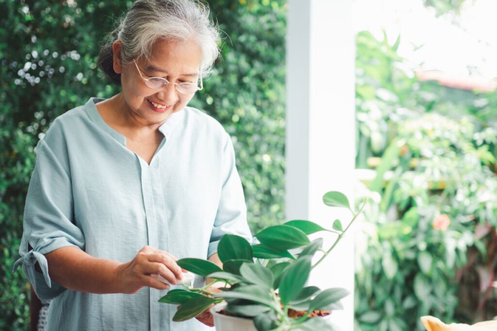 An older woman smiles gently while caring for a lush green houseplant in a bright, peaceful garden setting.