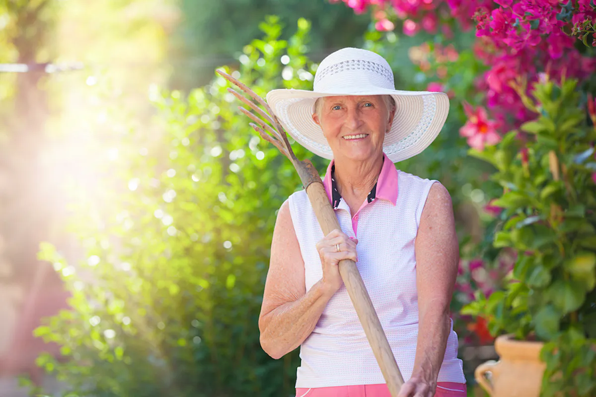 An older woman wearing a sunhat smiles while gardening outdoors in a vibrant summer garden filled with colorful flowers and bright sunlight.