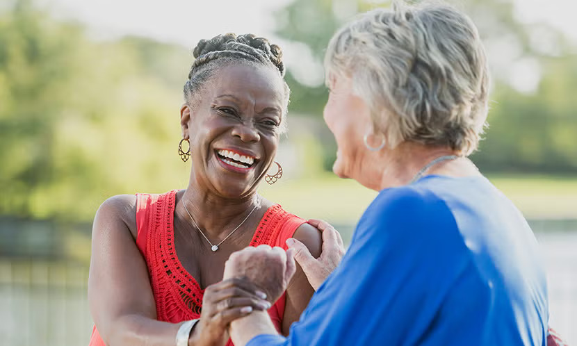 Two older women share a joyful moment outdoors, highlighting the warmth and connection that come from meaningful social relationships.