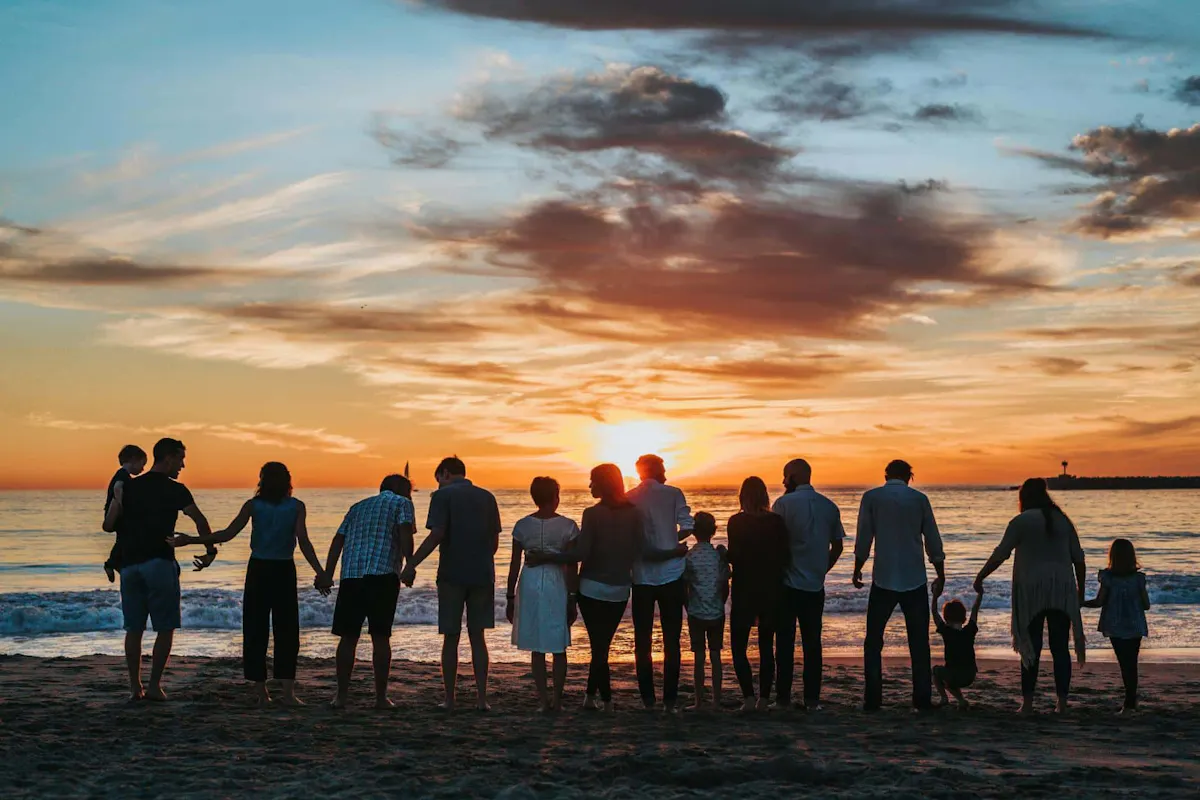 A multigenerational family stands together along the shoreline, holding hands and enjoying a beautiful sunset over the ocean.
