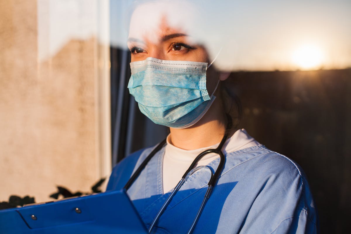 Young sad female caucasian UK US NHS EMS doctor carer looking through ICU window,fear uncertainty in eyes,wearing face mask gazing at sun,hope faith in overcoming Coronavirus COVID-19 pandemic crisis