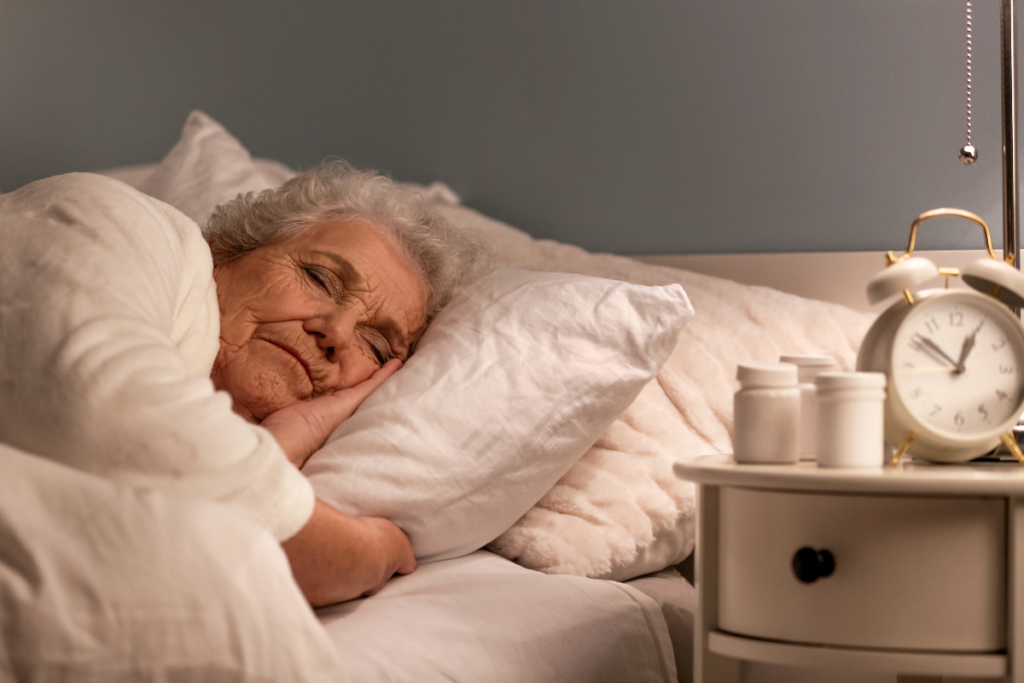 An older woman sleeps peacefully in bed beside a nightstand with an alarm clock and medication bottles.