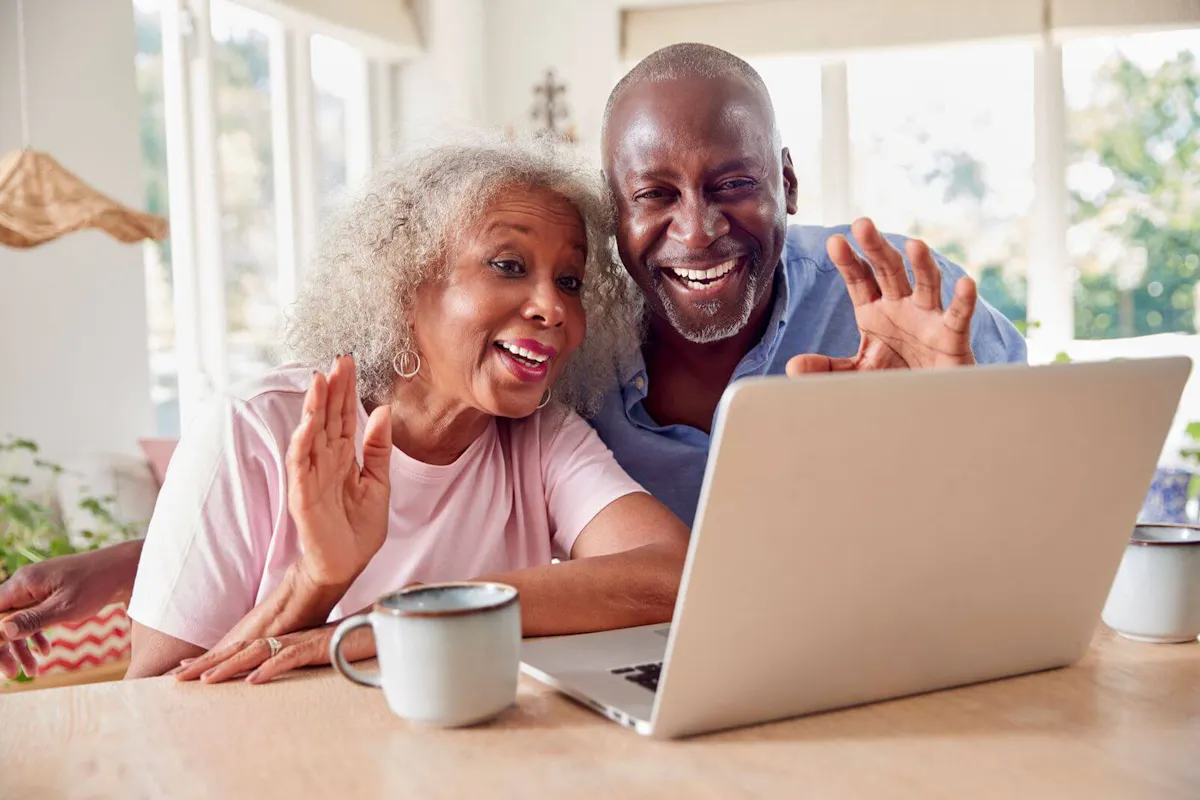 An older couple smiles and waves during a video call on a laptop, showcasing how technology helps seniors stay connected with loved ones.