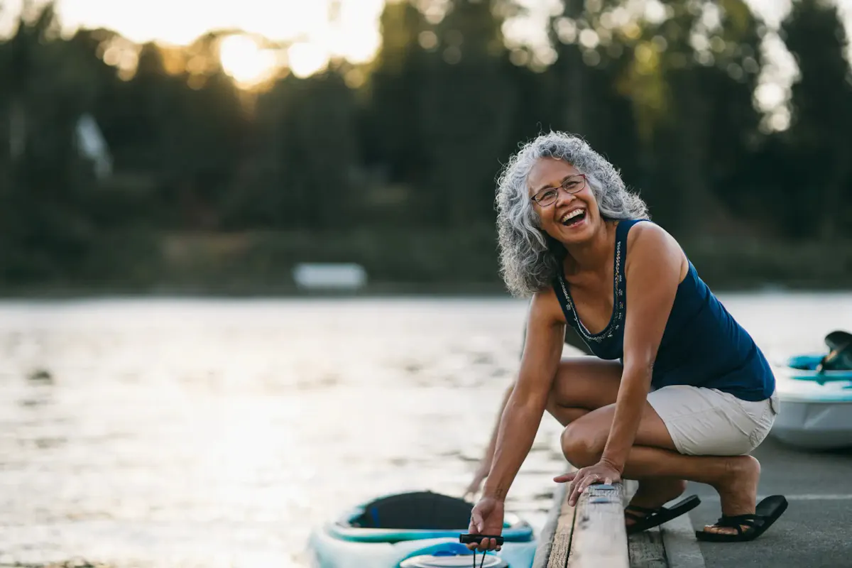 A smiling older woman enjoys a peaceful summer evening by the water, preparing for a relaxing kayaking adventure.