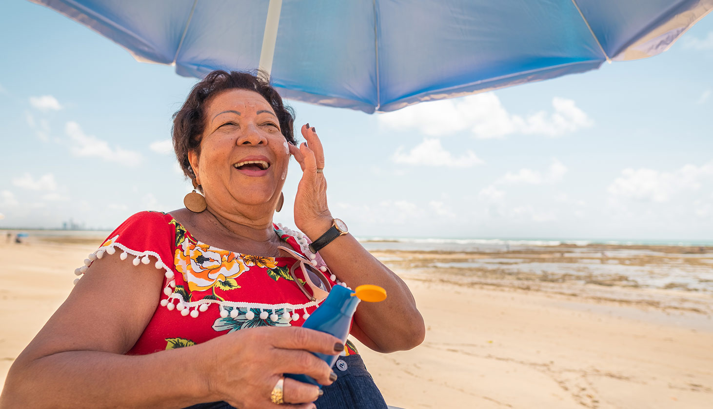 An older woman smiles while applying sunscreen under a beach umbrella on a sunny day by the ocean.