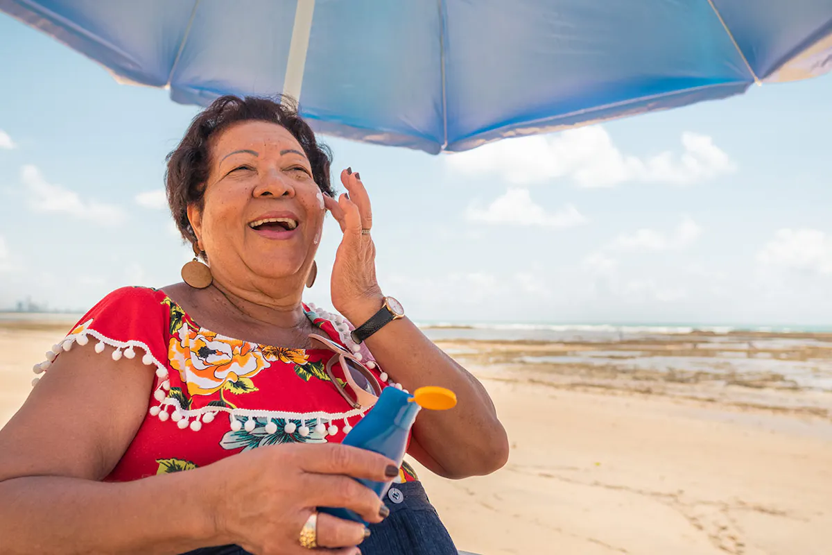 An older woman smiles while applying sunscreen under a beach umbrella on a sunny day by the ocean.