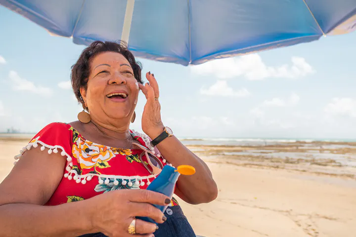 An older woman smiles while applying sunscreen under a beach umbrella on a sunny day by the ocean.