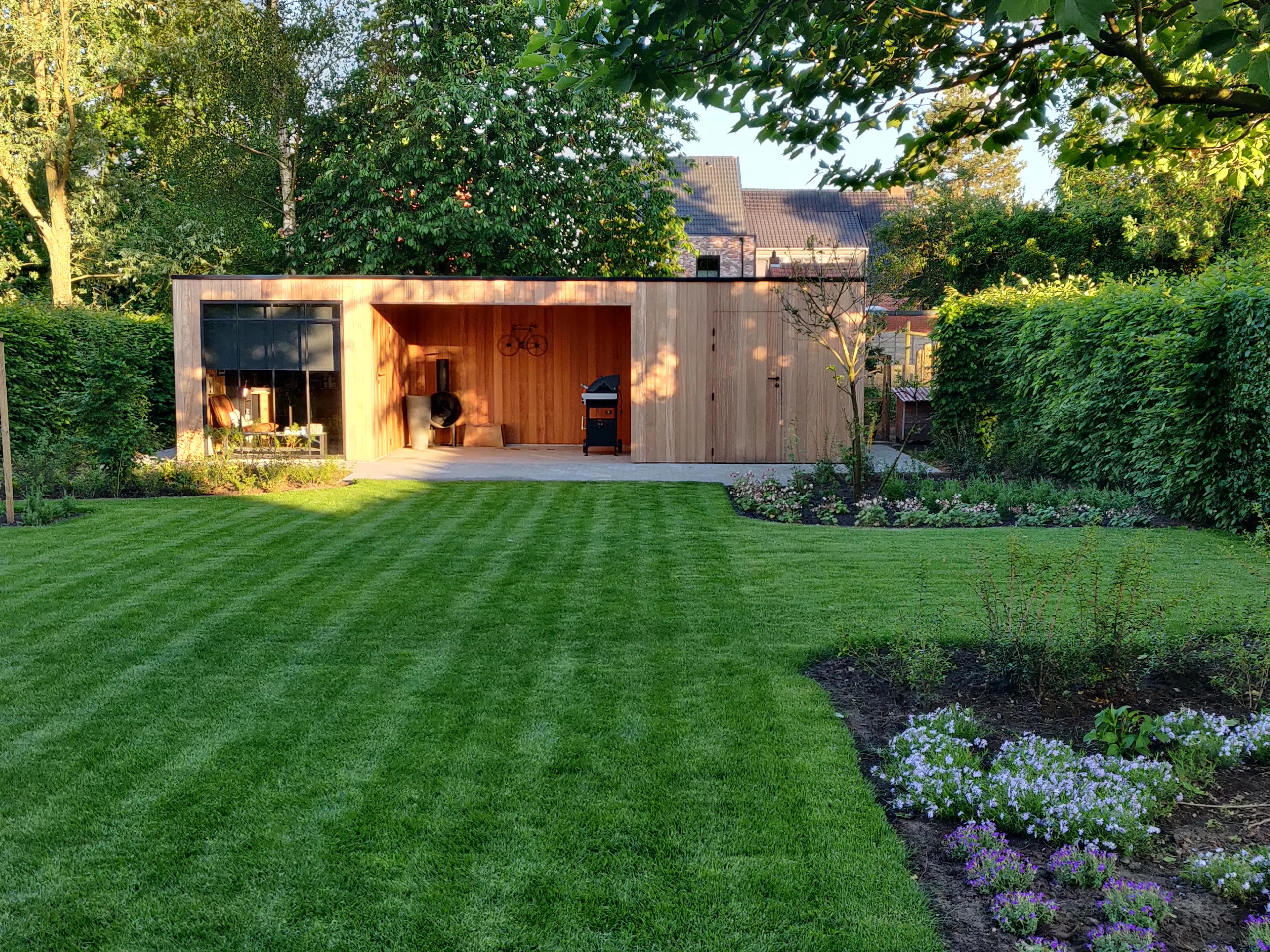 Moderne houten tuinberging met overdekte relaxruimte, plat dak, omgeven door een ruime groene tuin.