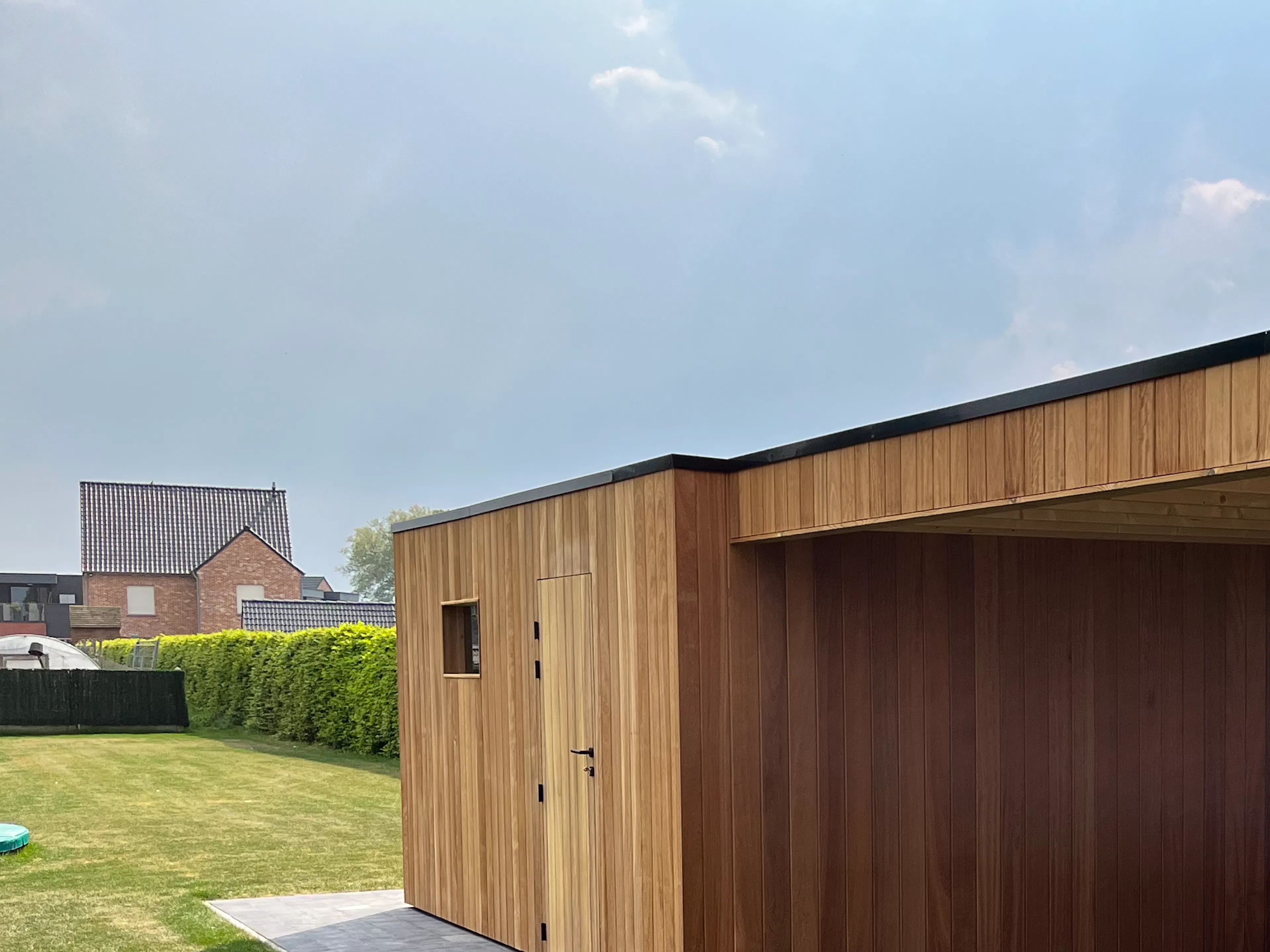 Moderne houten tuinberging met carport, Fraké thermohout, gelegen in een groene tuin.