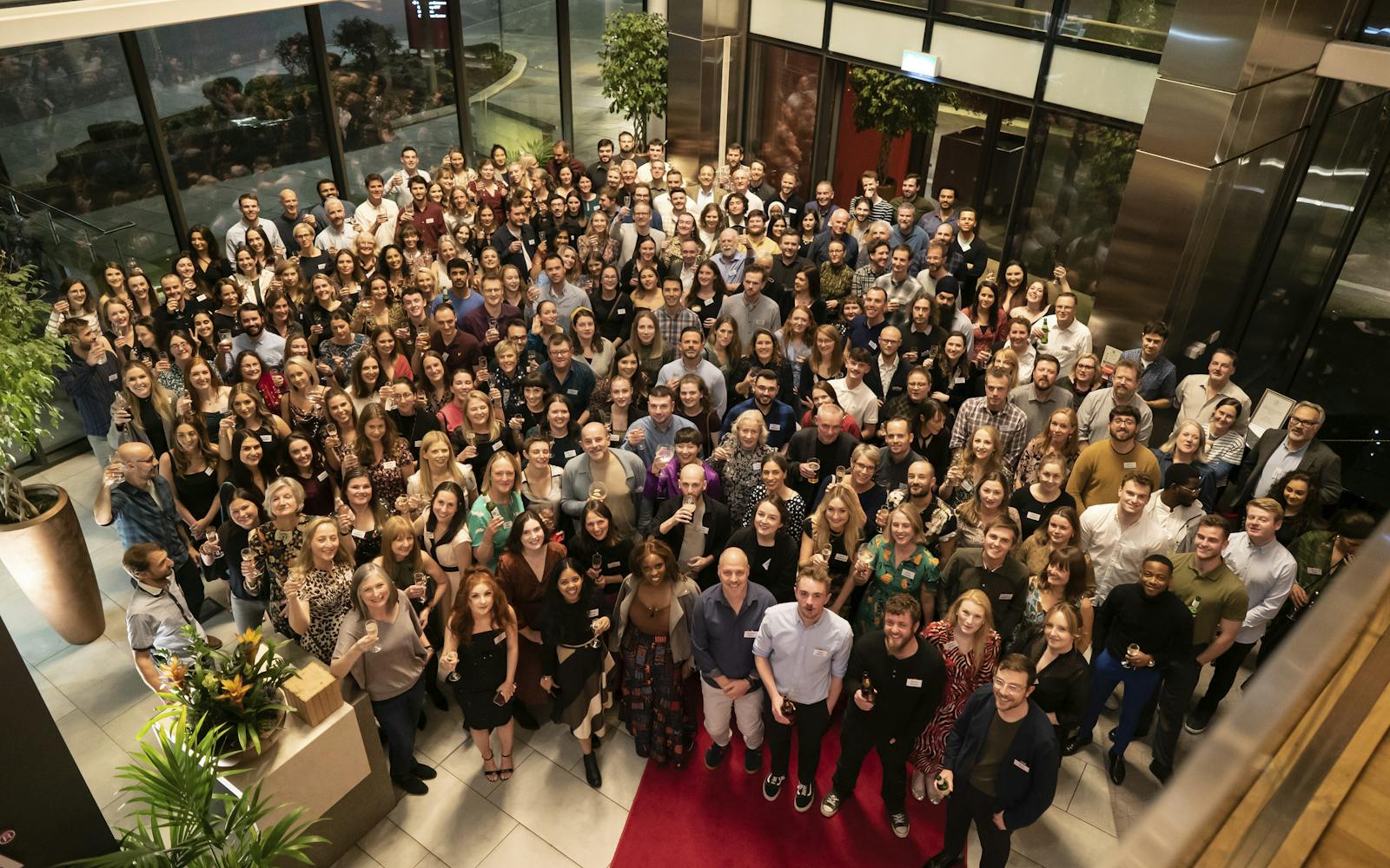 A large group of people gathered at a corporate event, smiling and holding drinks, inside a modern, well-lit venue with large glass windows and indoor plants. The diverse group includes men and women, all dressed in business casual attire, standing on a red carpet and looking up at the camera.