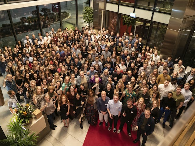 A large group of people gathered at a corporate event, smiling and holding drinks, inside a modern, well-lit venue with large glass windows and indoor plants. The diverse group includes men and women, all dressed in business casual attire, standing on a red carpet and looking up at the camera.