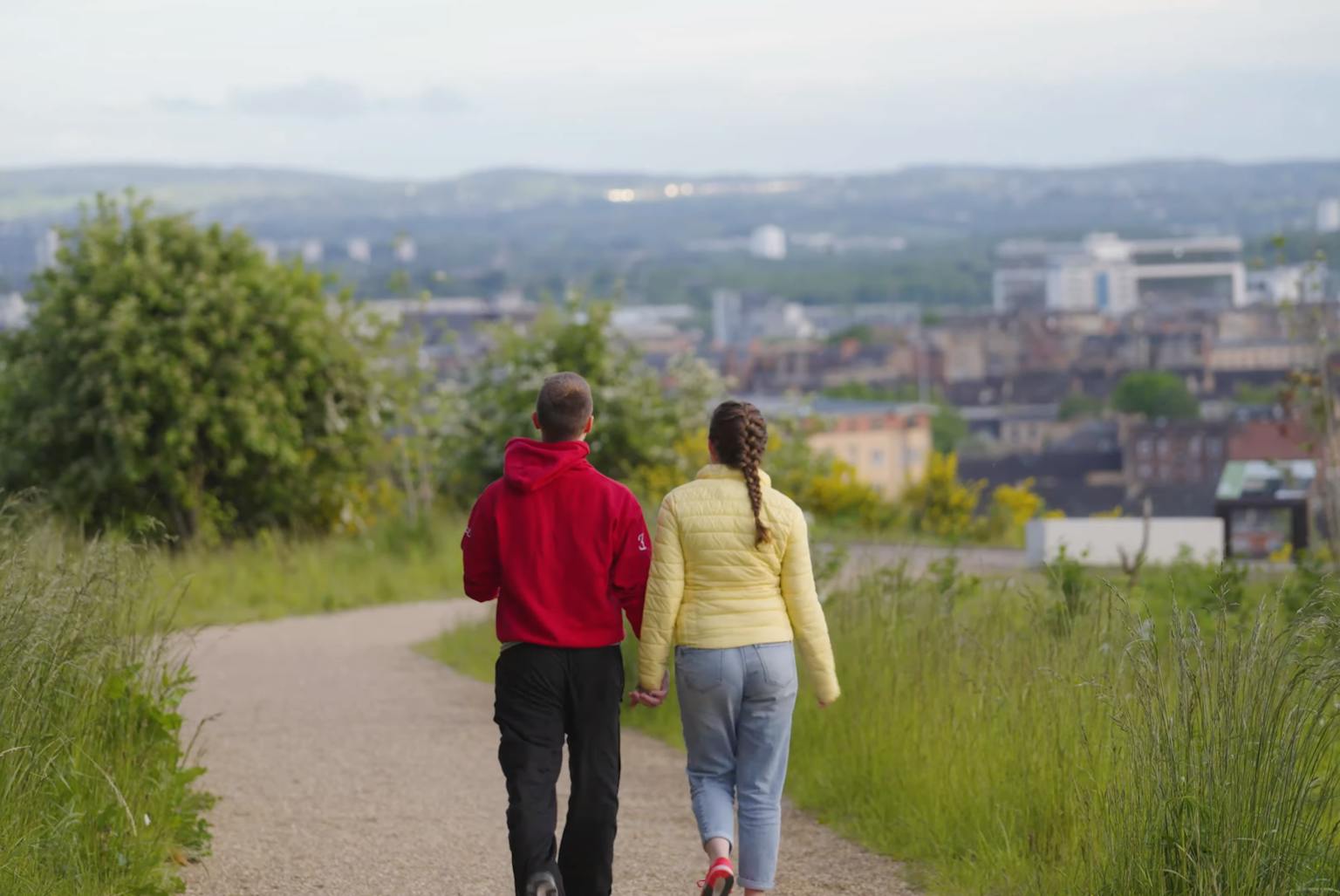 A couple holding hands and walking along a path surrounded by grass. The city is in the background