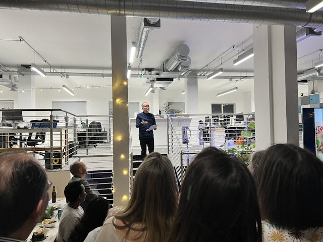 A man standing on a small indoor platform is giving a speech to a seated and standing audience in a modern office space, decorated with string lights.