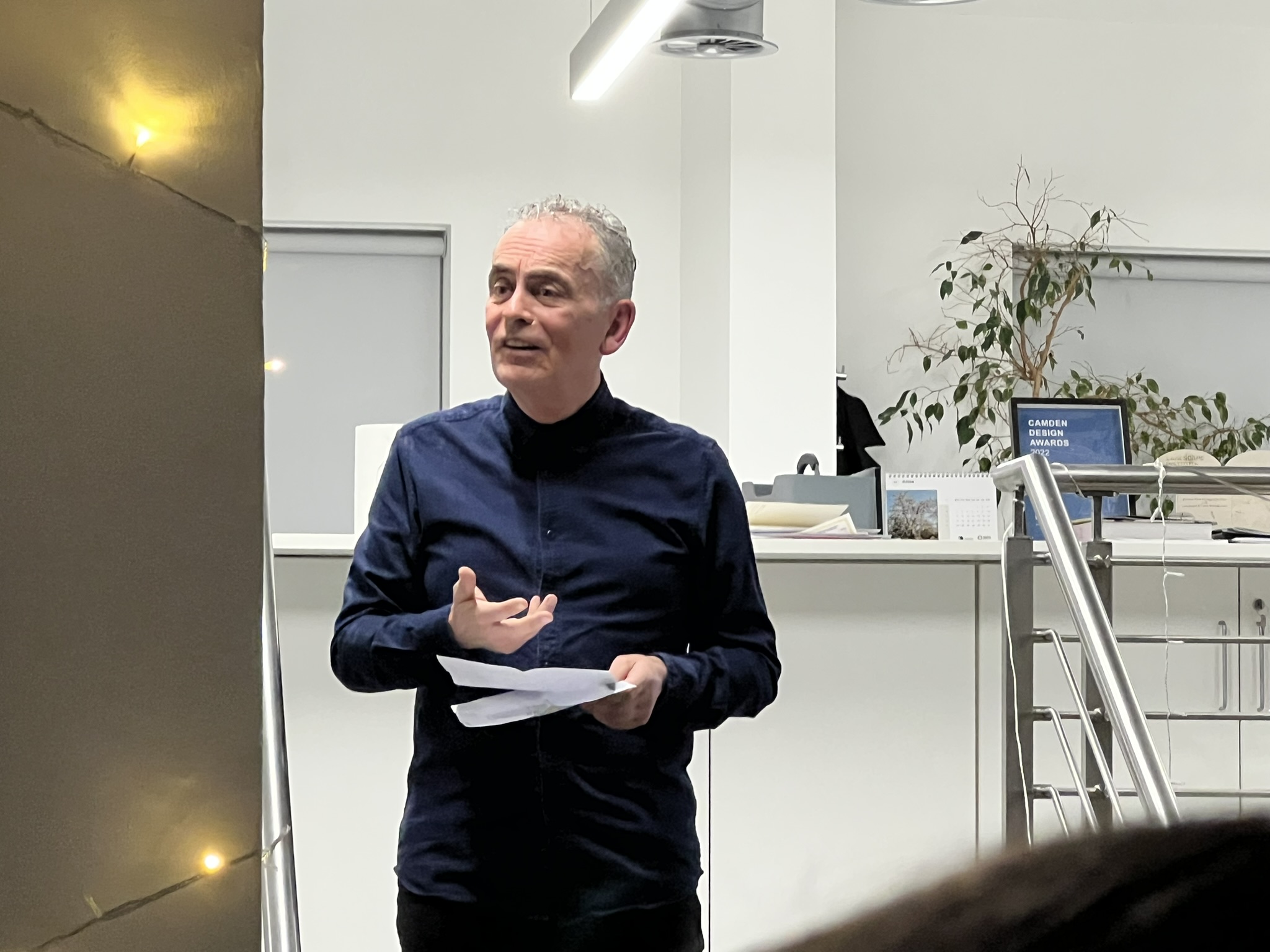 A person with short grey hair wearing a dark blue button-up shirt standing in an office space, gesturing with one hand while holding papers in the other. There's a potted plant and a desk with a calendar visible in the background.