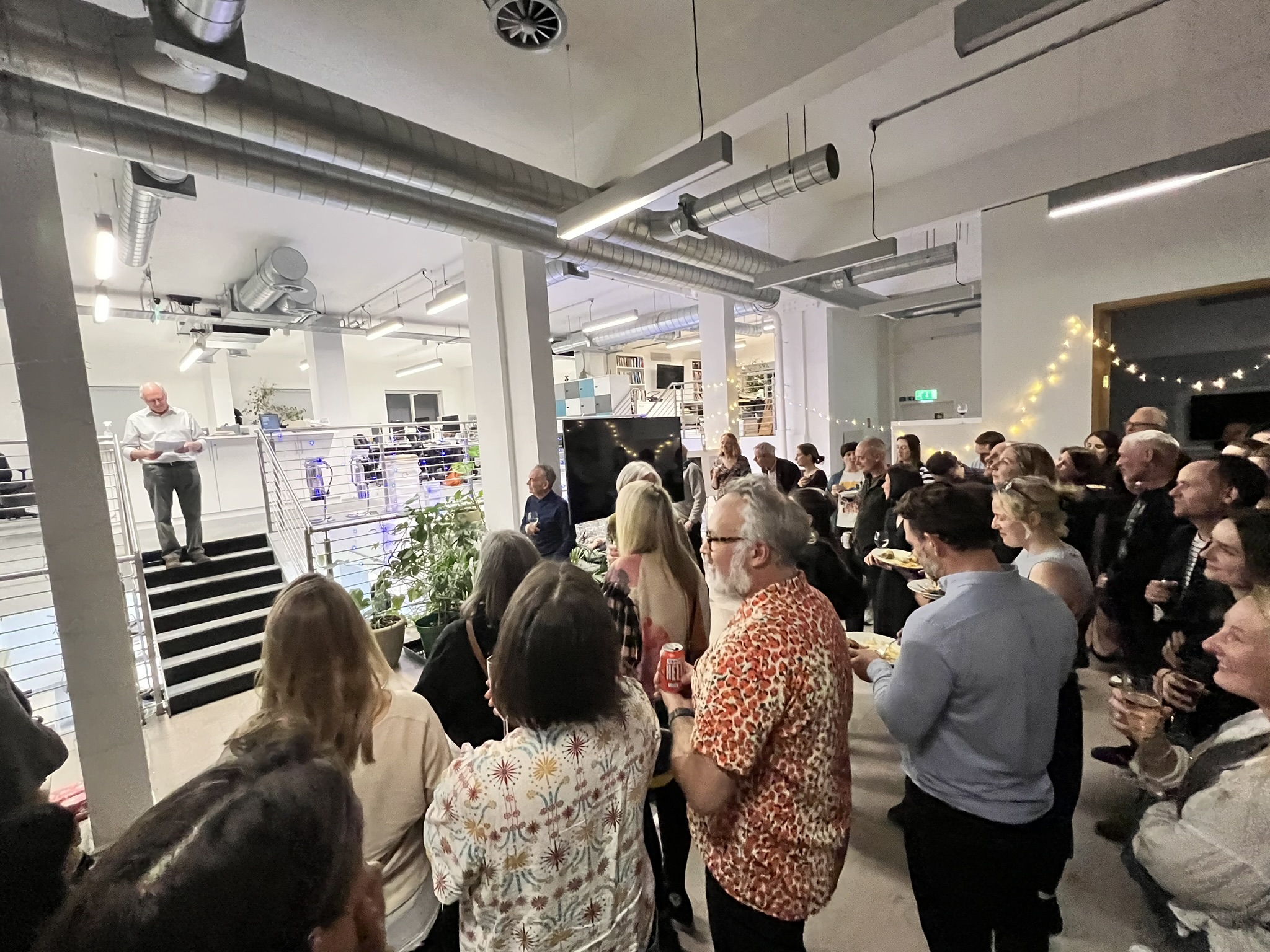 A person with white hair in light clothing standing on elevated stairs or platform addressing a crowd of people gathered below in an open office or industrial space with exposed ductwork. The audience members are holding drinks and appear to be at a social gathering or reception event.