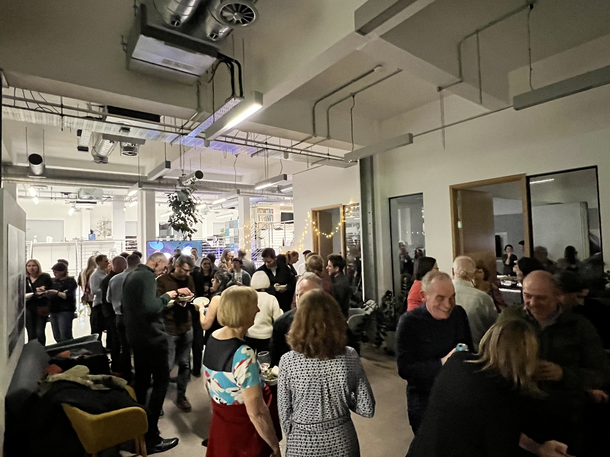A crowded indoor gathering or reception in an office space with exposed ceiling ductwork. People are mingling, holding drinks and plates, with string lights visible in the background. The industrial-style space features white walls, open areas, and appears to be hosting a social or networking event.