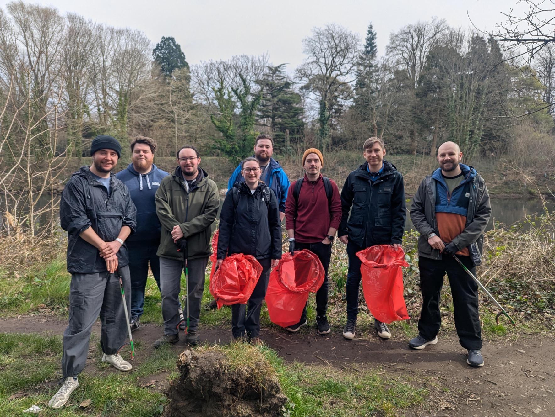 A group of eight volunteers standing outdoors in a wooded area, holding litter pickers and red rubbish bags, smiling at the camera.