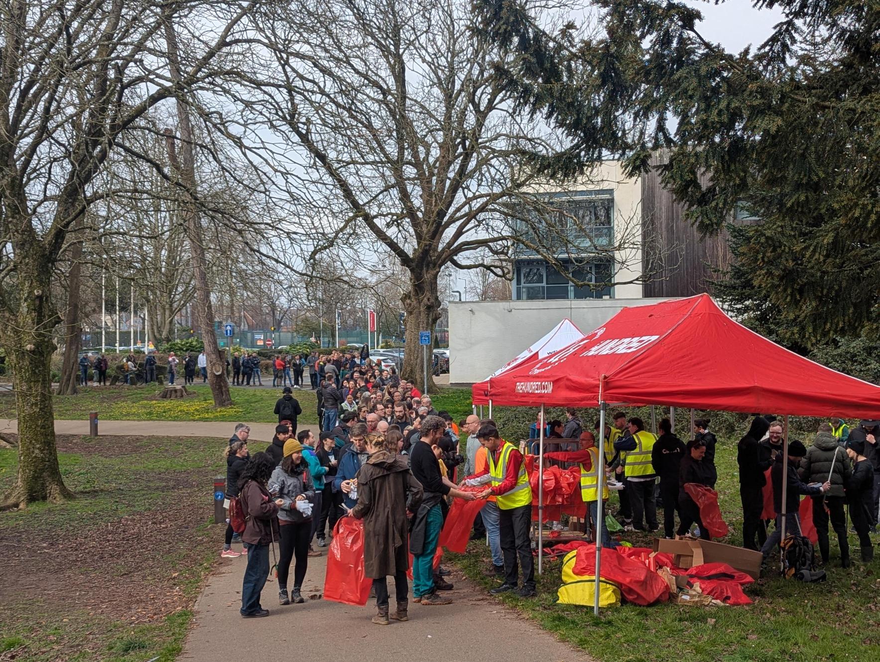 A busy outdoor scene with people lining up near red tents for a community cleanup event, surrounded by trees.