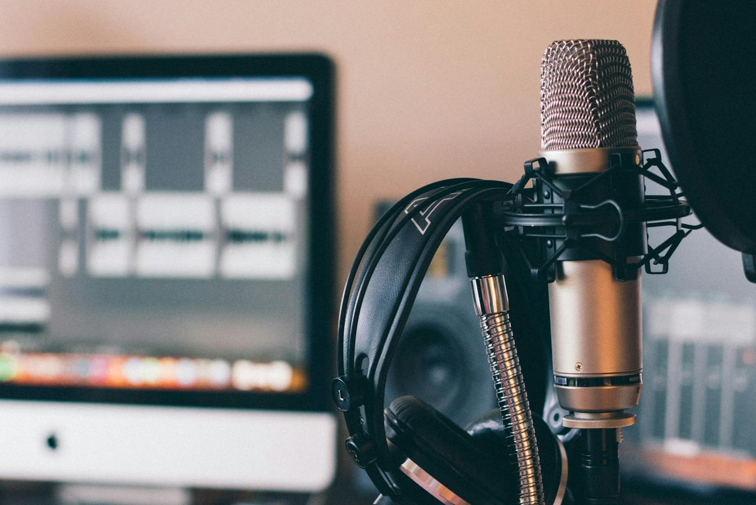 Professional condenser microphone with headphones next to a computer monitor in a home studio setup.