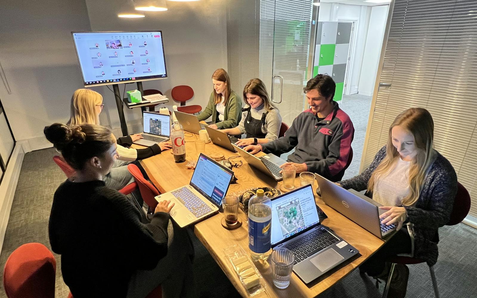 Six LUC colleagues collaborating during the 2024 GIS Day Mapathon. They are mapping Mozambique's Alto Molocue Province to support humanitarian aid efforts. A monitor displays mapping progress, highlighting their collective contribution to the Missing Maps initiative.