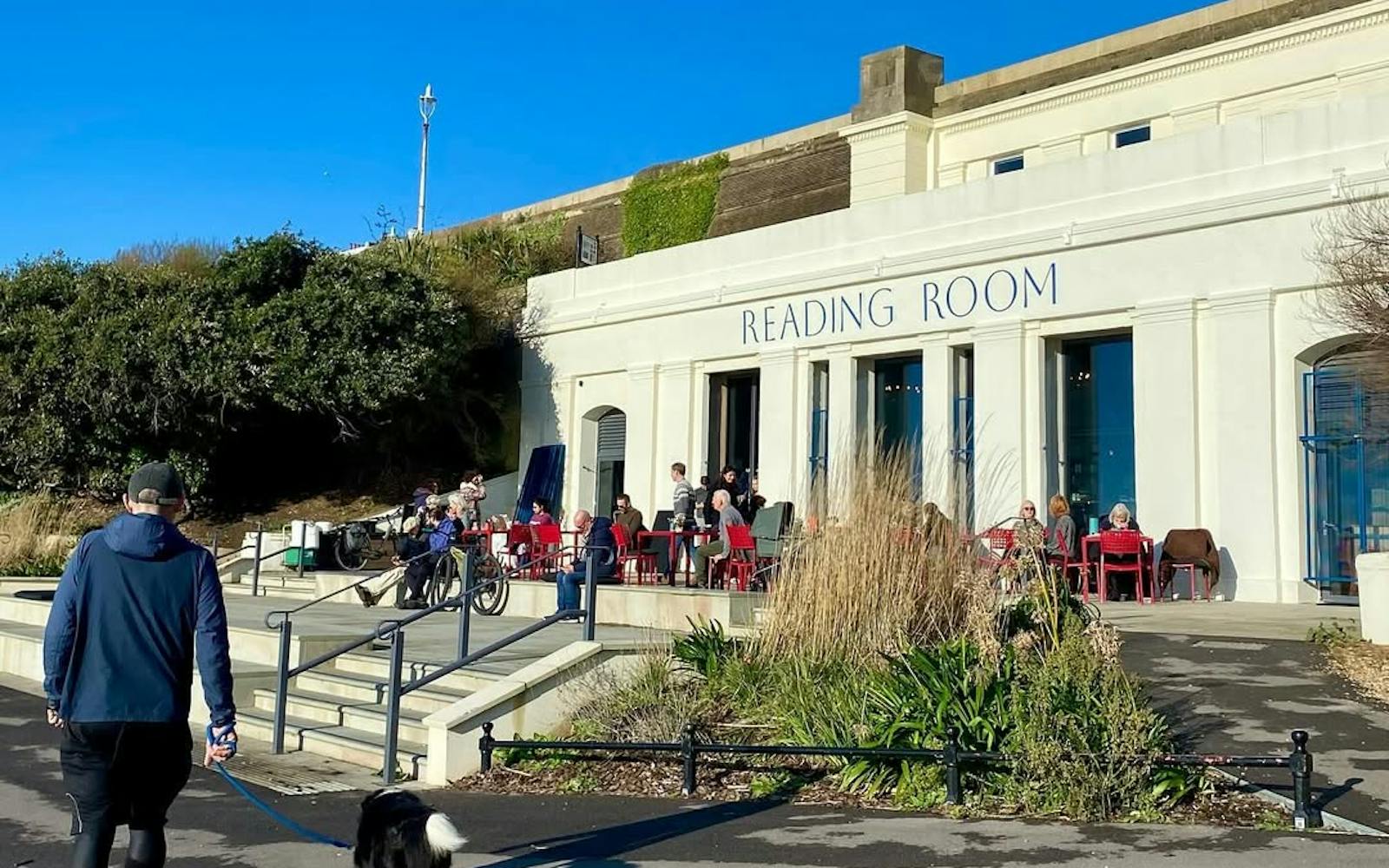The image shows the exterior of a building called the "Reading Room", with people sitting outside at tables. The building has an ornate, historic architecture style with a white facade and red roofs. Plants and landscaping surround the building. A person walking a dog can be seen in the foreground.