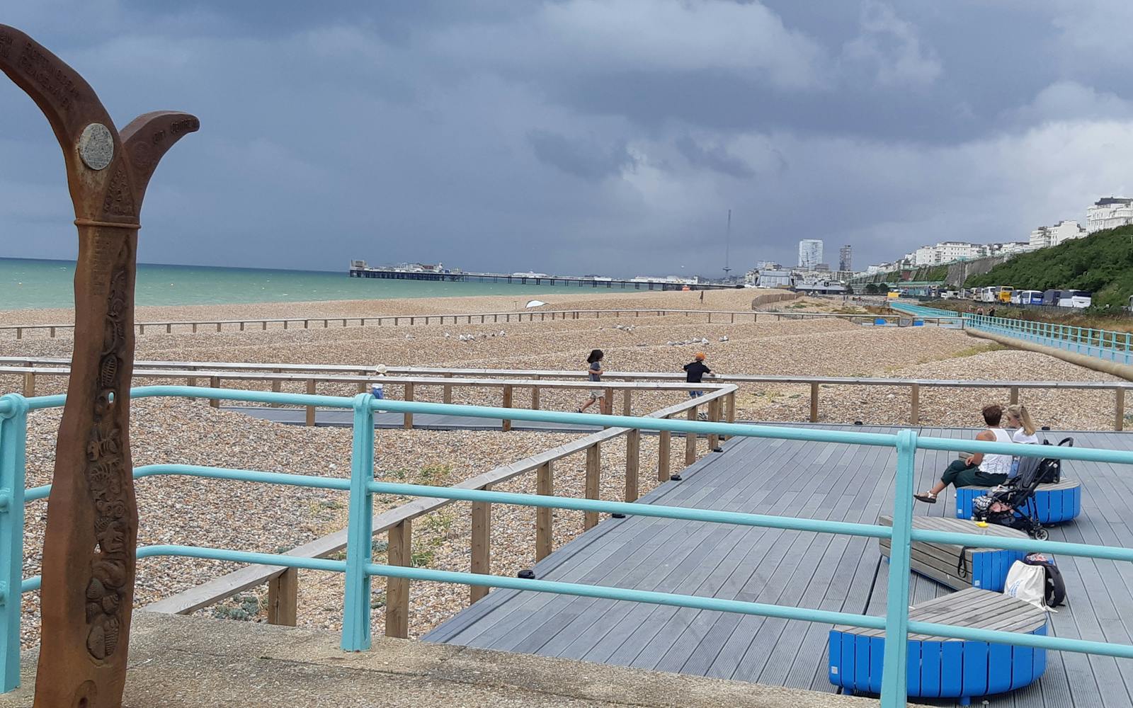 The image shows a scenic beach scene in what appears to be a coastal town. In the foreground, there is a wooden deck with benches and luggage, while the beach is visible in the background. The beach is covered in pebbles and there are some people walking on it. In the distance, there is a pier and a skyline of buildings. The sky is overcast with some clouds. The overall scene conveys a sense of a relaxing seaside location.