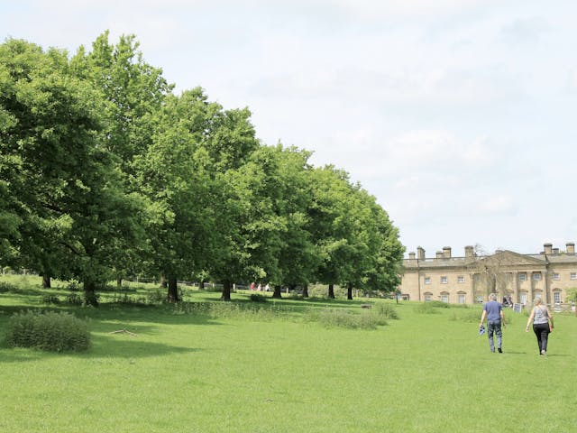 Two people walking across a field lined with trees towards a stately home