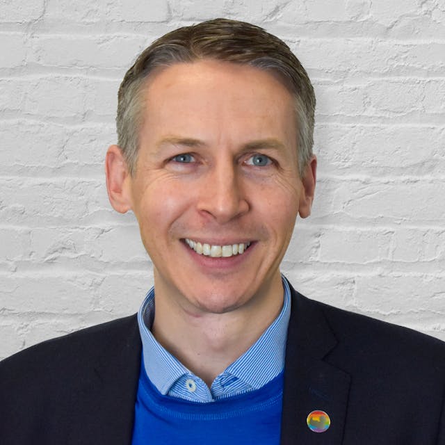 A smiling man with short, neatly styled brown hair and blue eyes stands in front of a white brick wall. He is wearing a black blazer over a blue sweater and a light blue collared shirt. A small round pin with a colourful design is attached to his blazer.