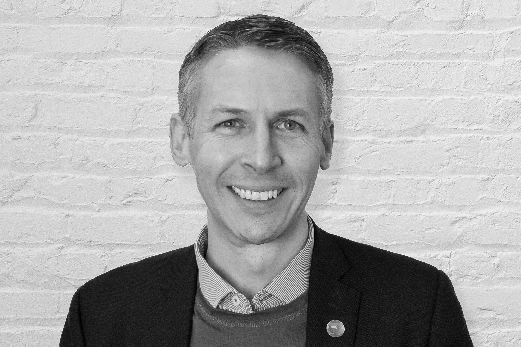 A smiling man with short, neatly styled brown hair and blue eyes stands in front of a white brick wall. He is wearing a black blazer over a blue sweater and a light blue collared shirt. A small round pin with a colourful design is attached to his blazer.