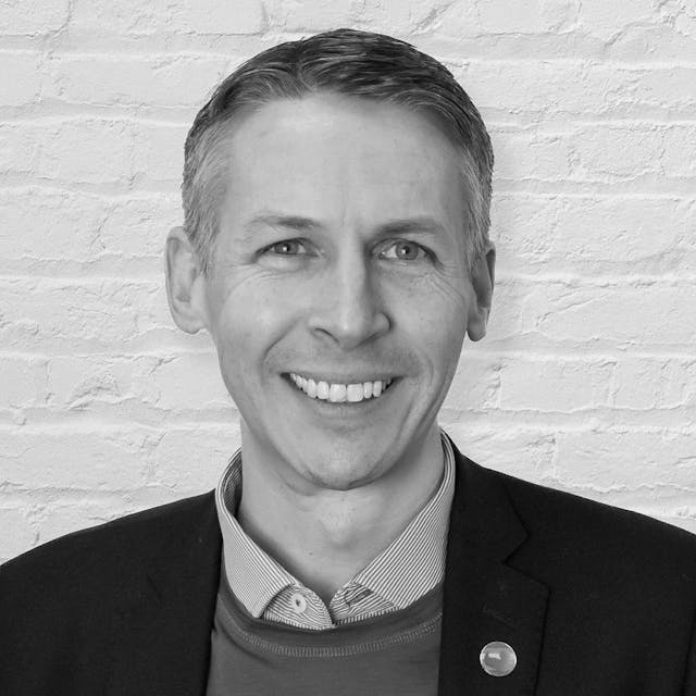 A smiling man with short, neatly styled brown hair and blue eyes stands in front of a white brick wall. He is wearing a black blazer over a blue sweater and a light blue collared shirt. A small round pin with a colourful design is attached to his blazer.