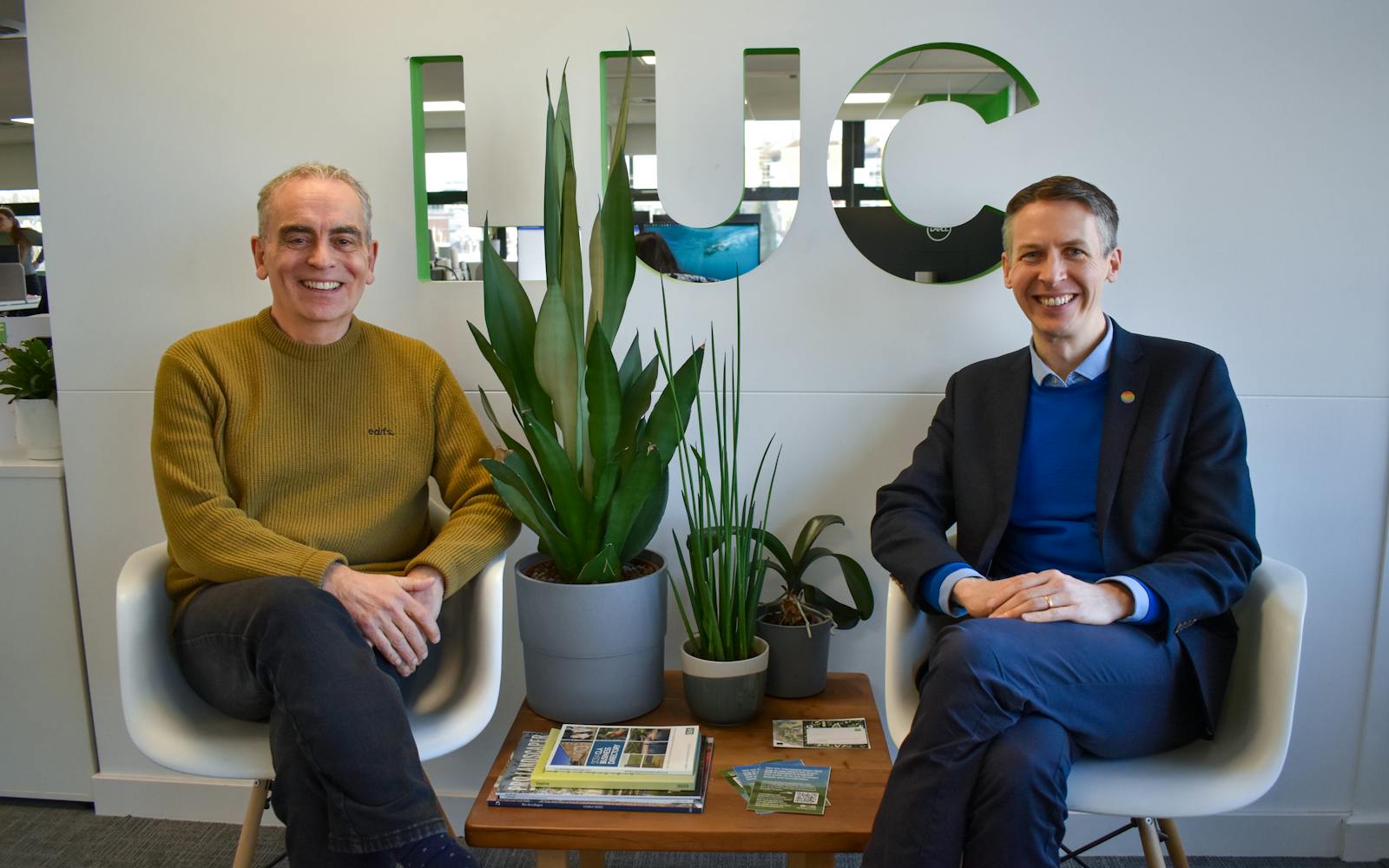 Two smiling men sit in white chairs in an office space with a modern design. Behind them, a wall features the letters "LUC" with mirrored cut-outs reflecting the office environment. A wooden table between them holds magazines, brochures, and small stacks of business cards. Several potted green plants are arranged on the table and floor. The man on the left wears a mustard-coloured jumper and dark trousers, while the man on the right wears a navy blazer over a blue sweater and collared shirt, with a colourful round pin on his lapel.