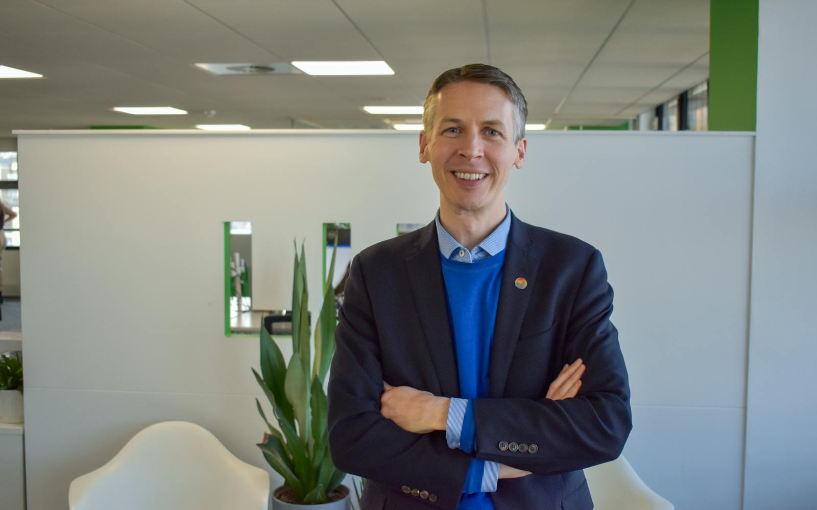 A smiling man with short brown hair stands with his arms crossed in a modern office. He wears a navy blazer over a bright blue sweater and a collared shirt, with a small colourful round pin on his lapel. Behind him, a white partition wall features mirrored cut-outs forming part of the "LUC" logo. A white chair and a potted green plant are visible in the background, along with office desks and windows.