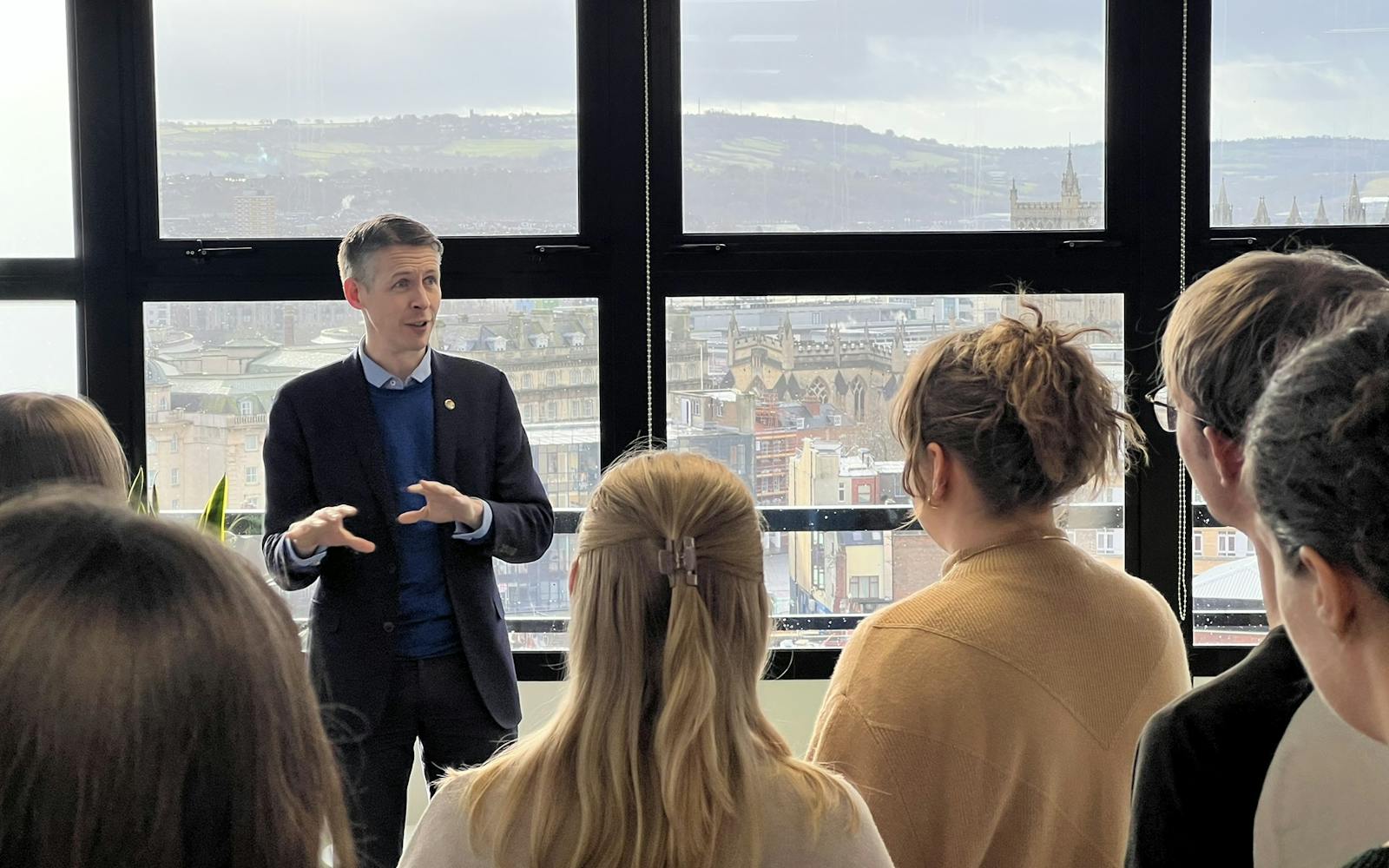 A man is giving a talk to a group of people in an office with a city view behind him.