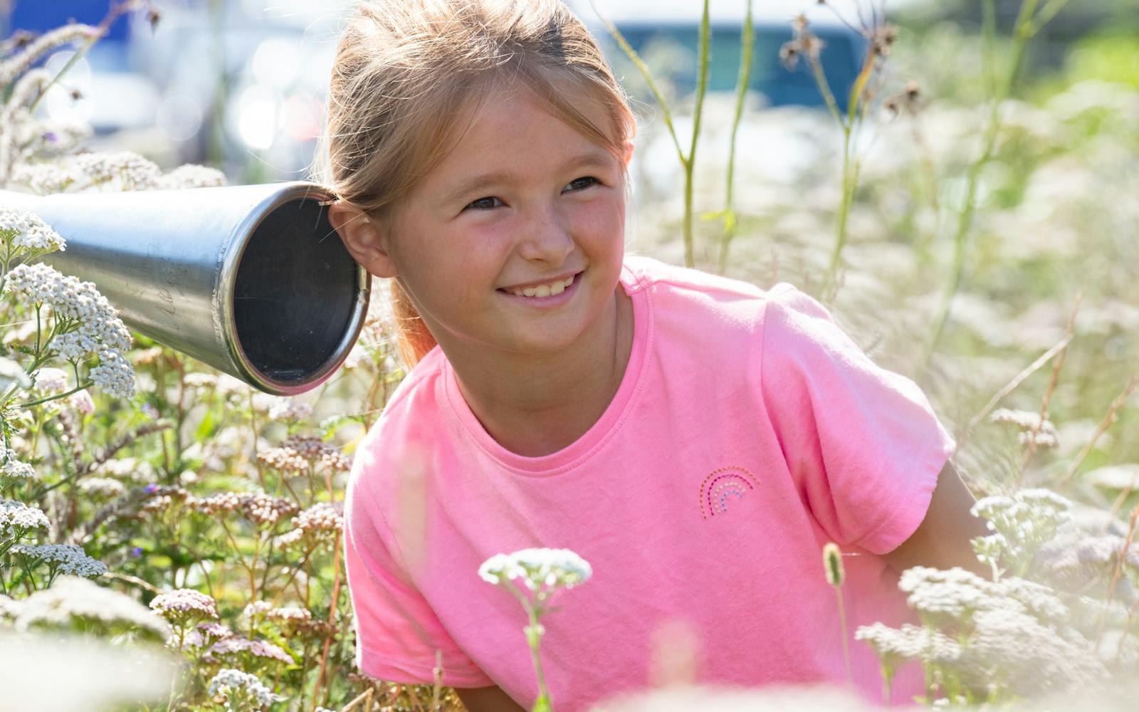 A child in a pink top surrounded by flowers with her ear to a tube