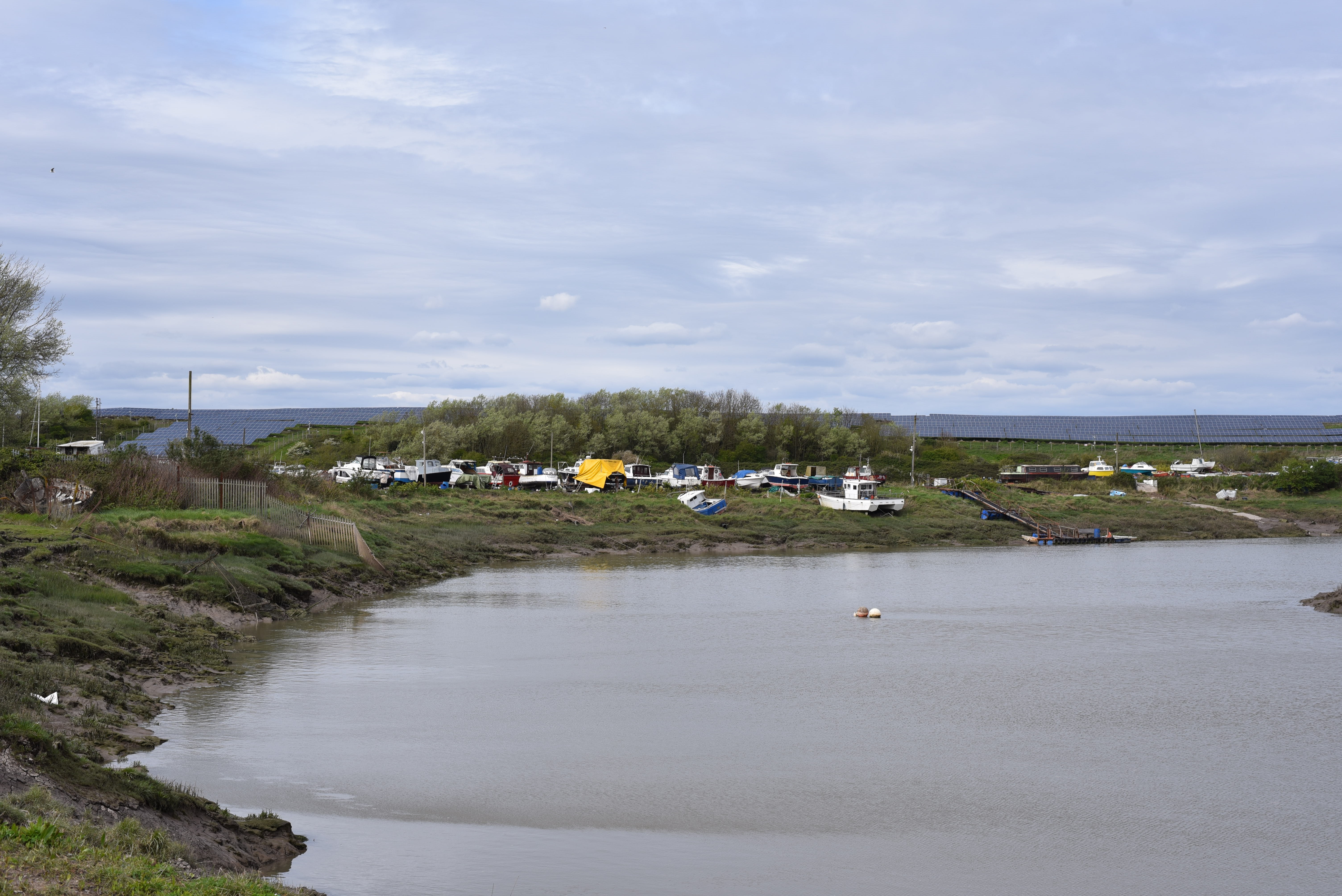 A muddy riverbank with boats and industrial structures in the background under a cloudy sky.