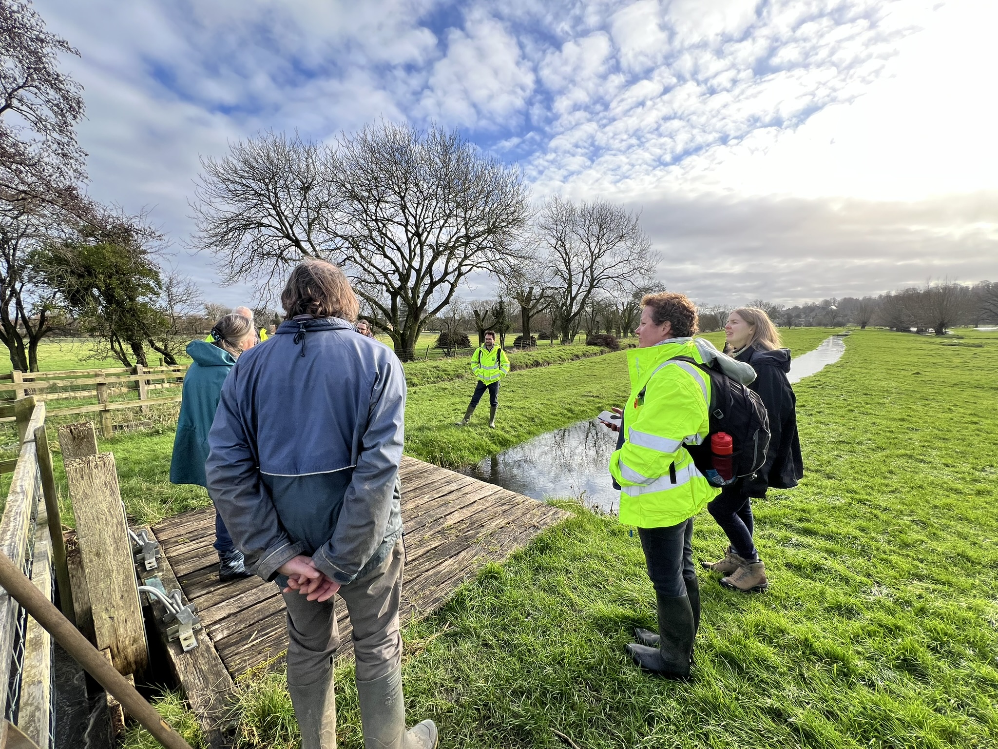 A group of conservationists and land management experts gather around a wooden structure in a meadow, discussing traditional water management techniques. Some wear high-visibility jackets, and one person holds a notepad.