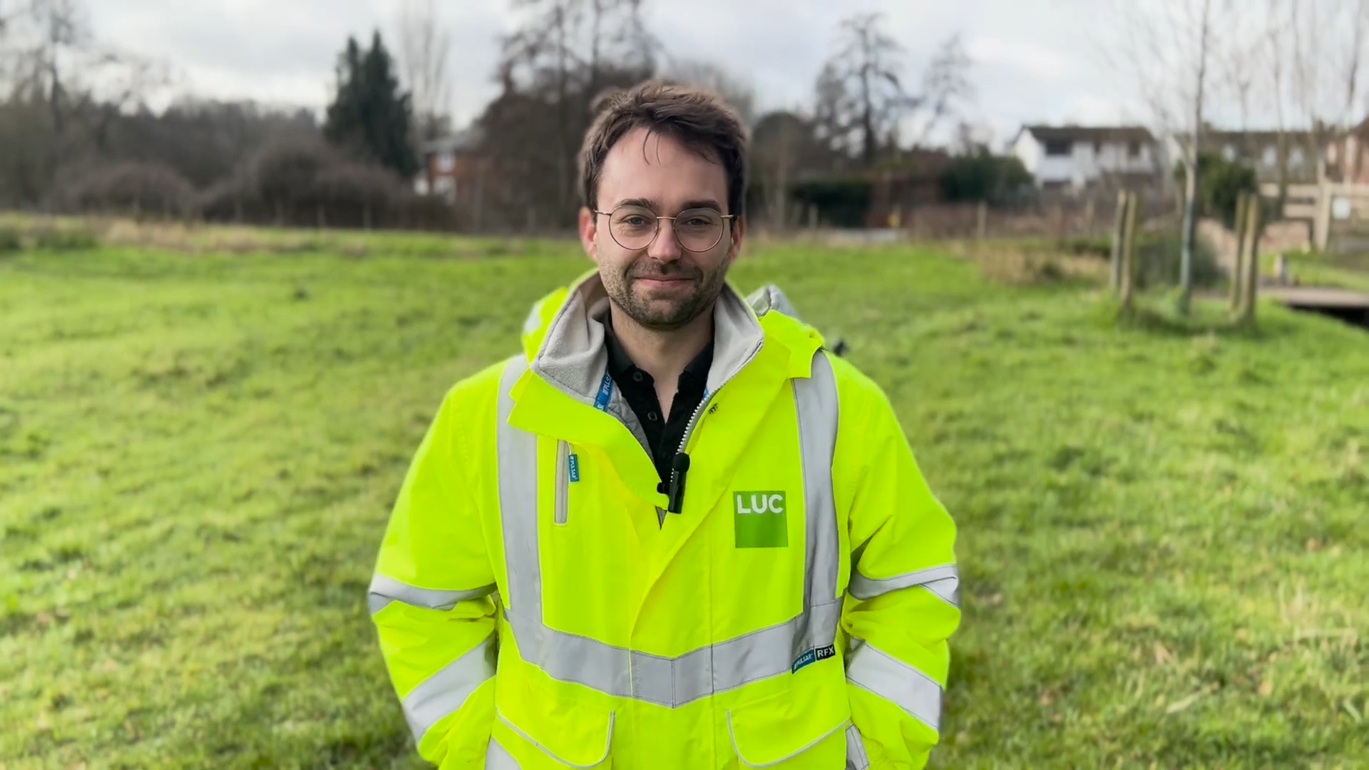 A man wearing a yellow hi-vis jacket standing in a green meadow and smiling at the camera.