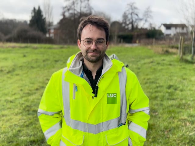 A man wearing a yellow hi-vis jacket standing in a green meadow and smiling at the camera.