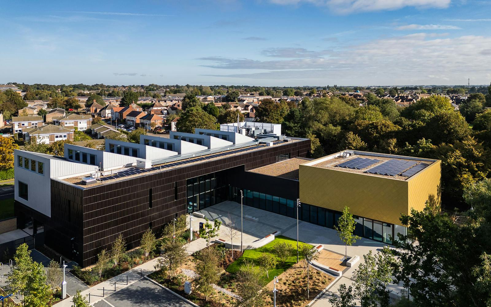 Modern public building with brown and yellow facades, solar panels on roof, surrounded by landscaped grounds and residential neighborhood.