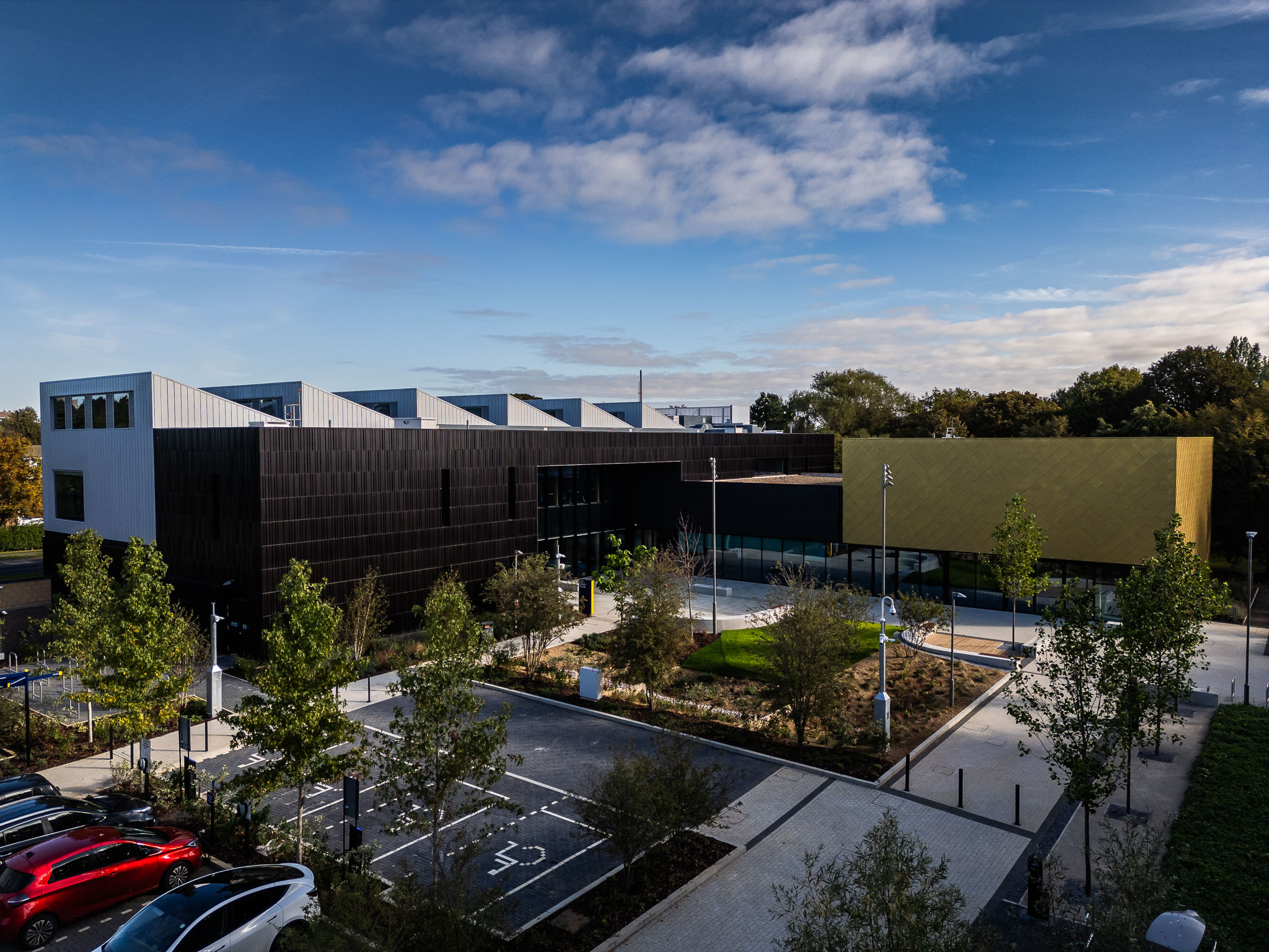 Modern building complex with white sawtooth roof, brown and gold facades, surrounded by landscaped grounds and parking area.