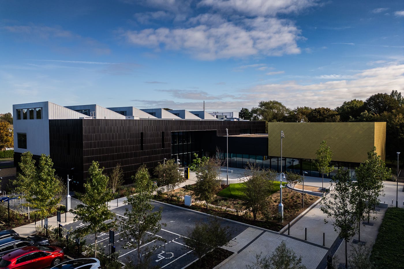 Modern building complex with white sawtooth roof, brown and gold facades, surrounded by landscaped grounds and parking area.