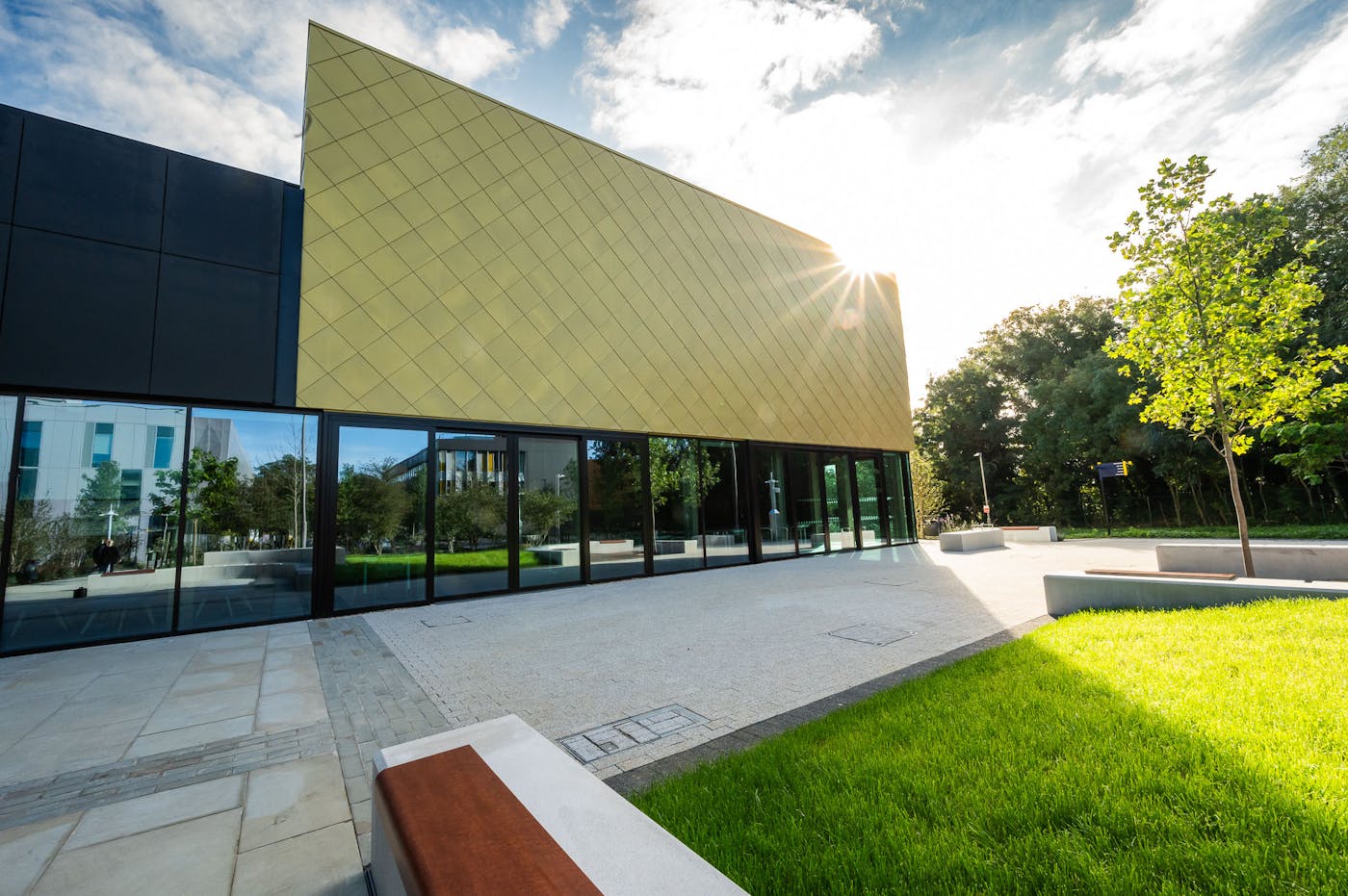 Modern building with gold geometric facade and glass walls, featuring sunlight reflection and landscaped outdoor area with grass and benches.