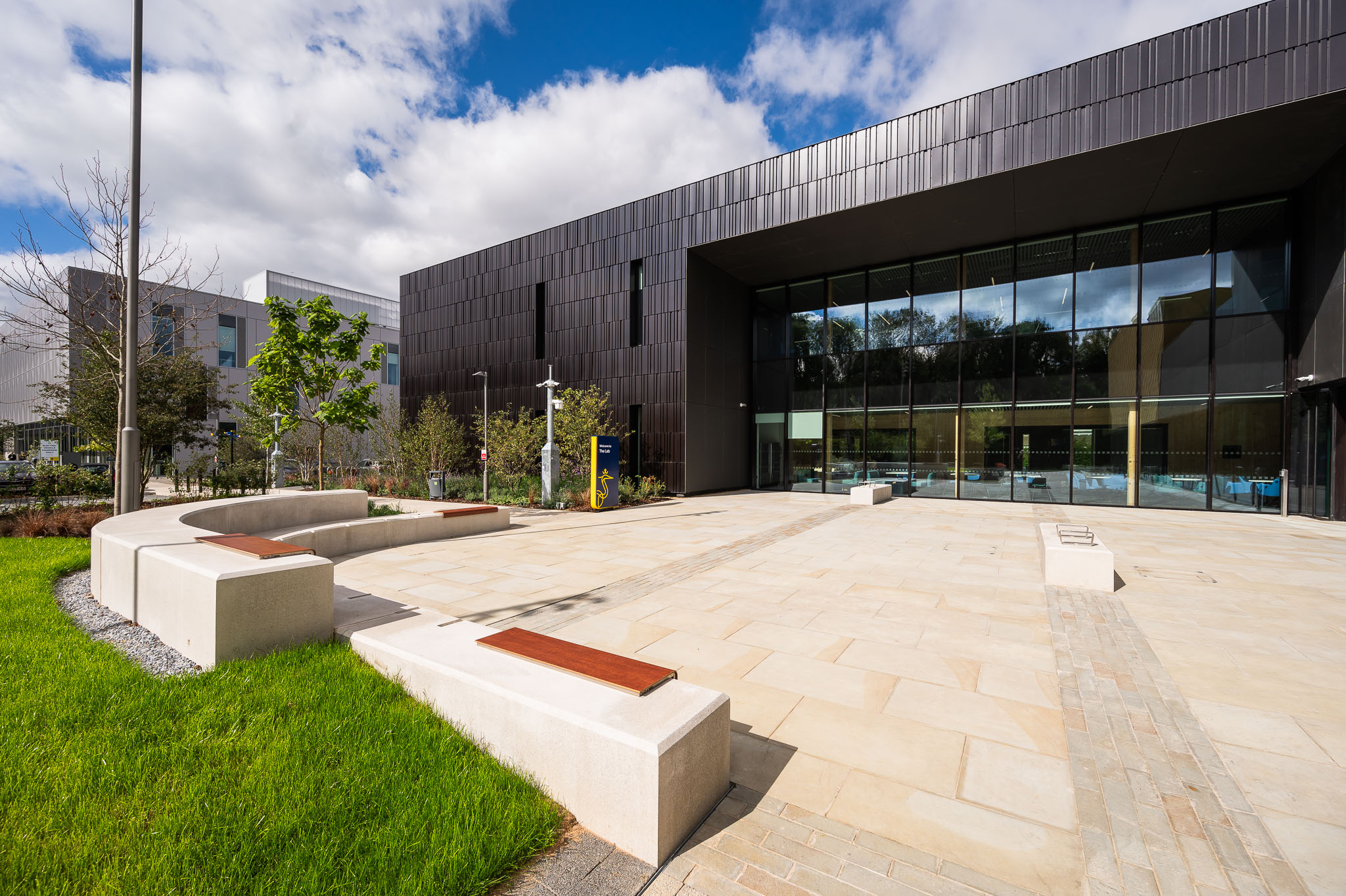Modern dark-clad building with glass entrance and outdoor plaza featuring concrete benches with wooden seating, surrounded by landscaped grounds.