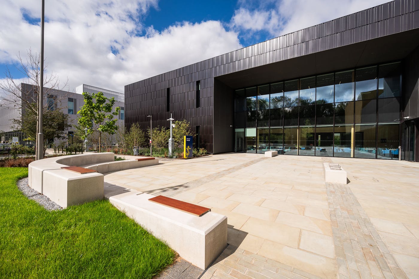 Modern dark-clad building with glass entrance and outdoor plaza featuring concrete benches with wooden seating, surrounded by landscaped grounds.