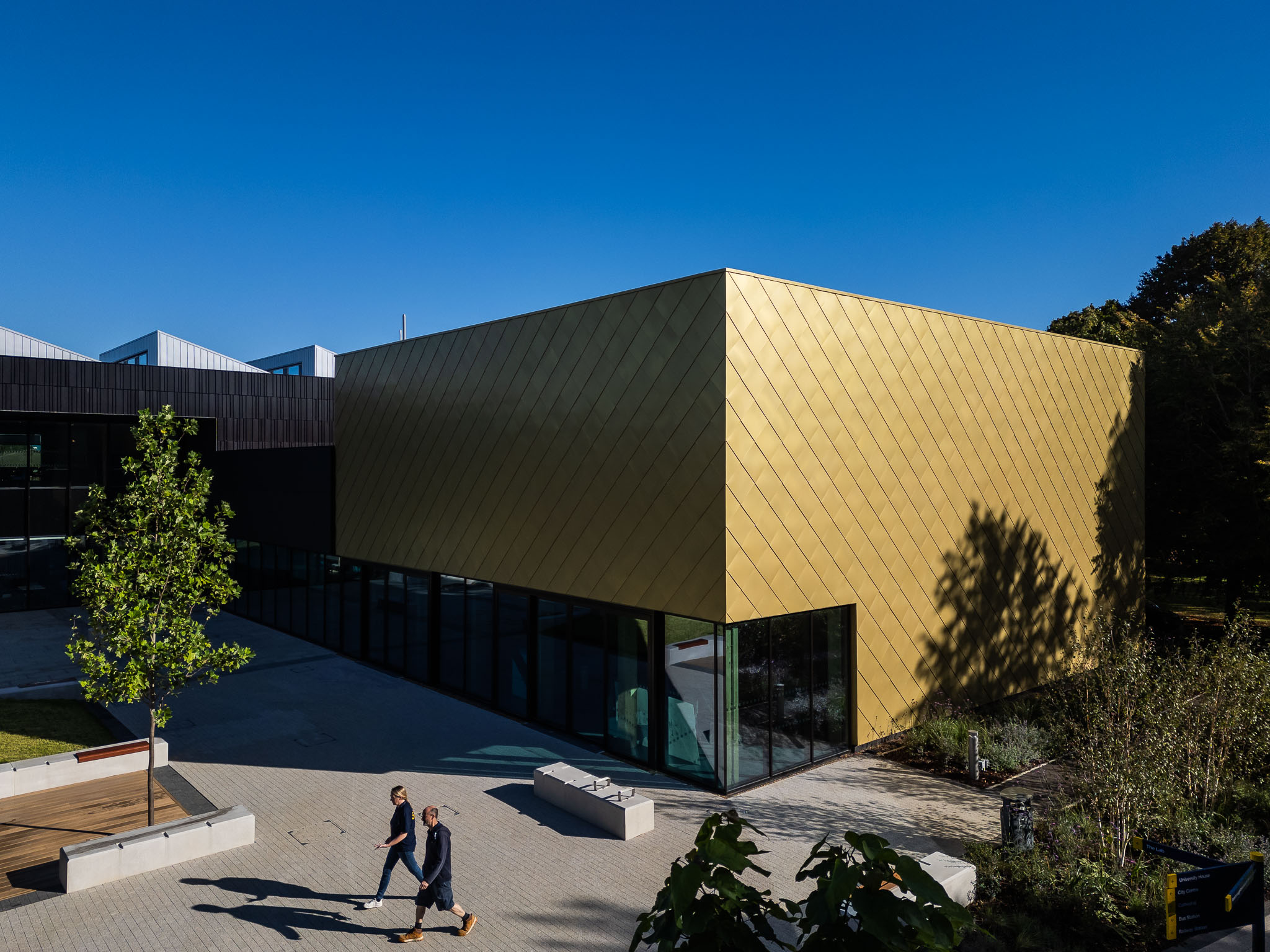 Modern building with gold geometric facade and dark sections, featuring glass walls and landscaped surroundings, with two people walking on the paved area below.