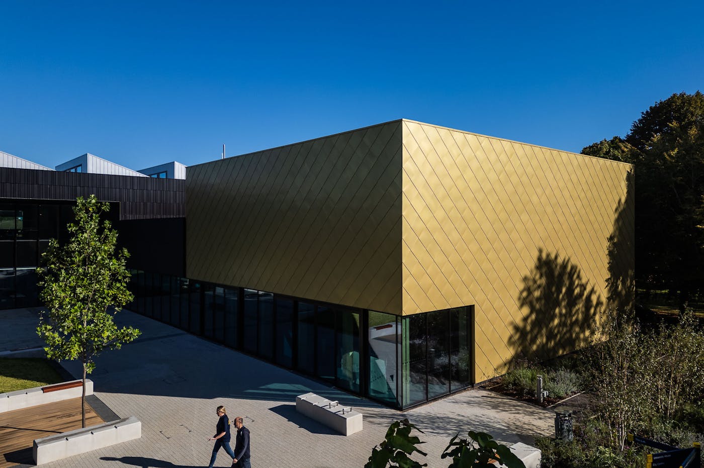 Modern building with gold geometric facade and dark sections, featuring glass walls and landscaped surroundings, with two people walking on the paved area below.