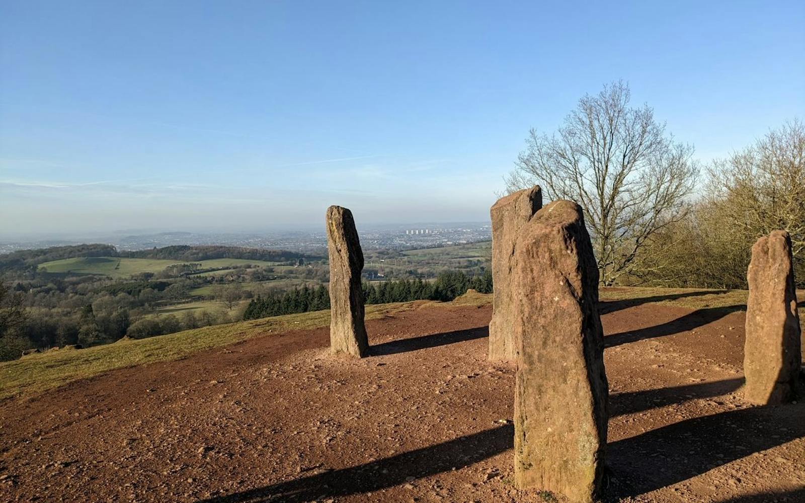 A group of standing stones on a hilltop overlooking a landscape of fields, trees, and a distant city under a clear blue sky.